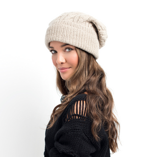 A young woman wearing a Cobain Slouch hat handmade in Nepal, beige knitted hat and a black sweater with a shoulder cut-out detail smiles faintly while glancing sideward.