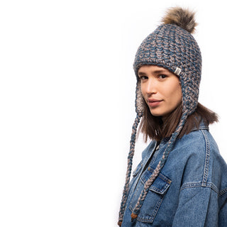 A person wearing the Chunky Knit Earflap Hat, featuring a pom-pom, stands against a white background. Dressed in a denim jacket, they are looking at the camera.