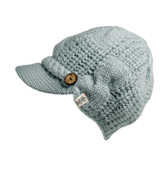 A Hand Knit Broadway Hat in light blue, crafted in Nepal, featuring a short brim and a wooden button accent on the side. It includes a white tag with a small logo. The hat is thick and textured like a cable-knit wool design, made to provide warmth.