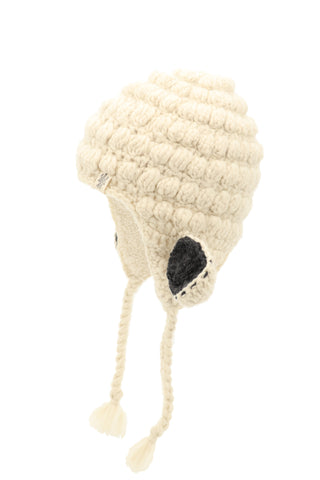 The Lamb Ears Hat is a cream-colored, hand-knitted beanie with ear flaps and braided tassels. It features textured, bubble-like stitching and two round, black patches on the ear flaps.