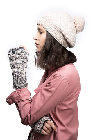 A woman wearing a beige knit beanie and a pink shirt holds up a handmade in Nepal merino wool glove, branded as Gotham Handwarmers, to her profile against a white background.productId.