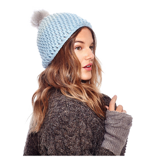 A woman wearing a blue Dimensions Crochet Beanie with Faux Fur Pom looks over her shoulder with a slight smile, wearing a warm sweater and fingerless gloves.