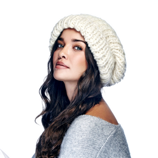 A woman with long dark hair wearing a handmade in Nepal Chunky Slouch w/ Fold white beanie and an off-the-shoulder light gray sweater. She is looking to the side with a thoughtful expression.