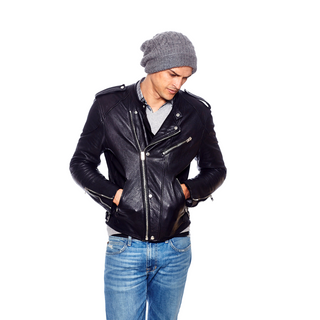 A man wearing a Cobain Slouch black leather jacket, jeans, and a gray beanie with fleece lining and 100% wool stands with his hands in his pockets looking downwards.