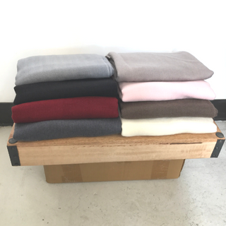 A stack of neatly folded sweaters made from Solid Pashmina Scarf wool in various colors placed on a wooden bench against a white wall.