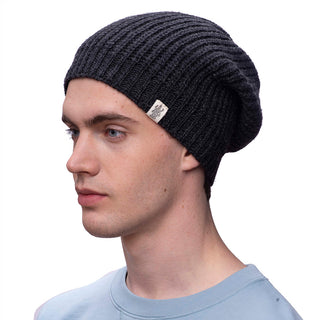 A person wearing a dark Cardigan knit slouch beanie, handmade in Nepal, and a light blue shirt is looking to the left against a plain white background.