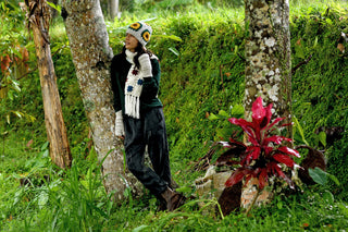 A person with long, dark hair stands in a green field wearing the Chunky Hat, a rust-colored scarf and mittens, and a dark green sweater, gazing slightly to the side.