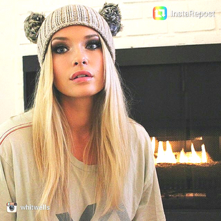 A woman wearing a Double Pom Beanie sits in front of a fireplace.