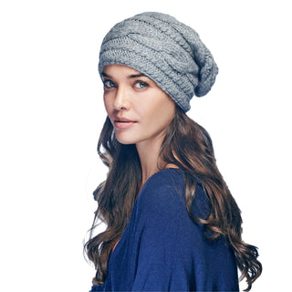 A woman with long brown hair wears the Triple Braid Cable Slouch, a gray hand-knit winter hat, comfortably on her head as she faces slightly to the side in front of a plain white background, looking at the camera while wearing a navy blue sweater.