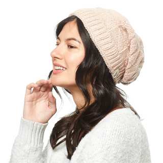 A person with long dark hair is shown in profile against a white background, looking ahead and smiling slightly while wearing the Wave Slouch, a light pink Merino wool hat, along with a white sweater.
