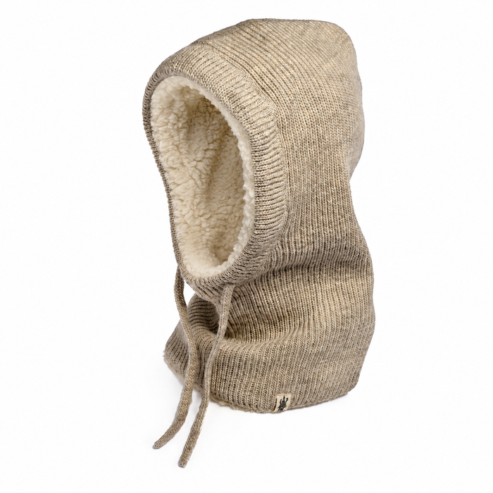 The Hand Knit Cozy Hood, a beige ribbed knit hooded balaclava featuring cream sherpa fleece lining and drawstrings, is displayed upright against a plain white background. A small woven tag accents the bottom edge of the hood.