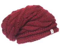 A deep red, chunky Hand Knit Triple Braid Cable Slouch hat featuring a classic cable pattern and a small white fabric tag on the brim, displayed against a white background.