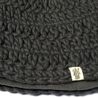 Close-up of the Hand Crochet Acorn Hat, showcasing its thick, dark gray, chunky knit texture reminiscent of a soft wool winter beanie. A small beige fabric tag is sewn onto the edge, featuring a printed design or logo.