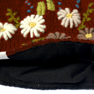 Close-up of the Hand Knit Bloom Hat—a brown, hand-knit wool hat adorned with embroidered white and yellow flowers and green leaves—showing its black fleece-lined interior and a white tag at the edge.