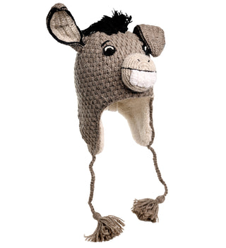 The Donkey Hat is hand-crocheted to resemble a donkey's head and includes a smiling face, floppy ears, black mane, and long tassels hanging from each side. This charming accessory is grey with white accents and appears to be made of wool.