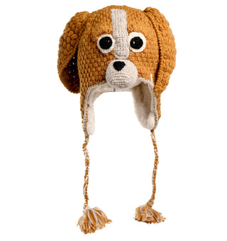 The Crochet Lucy Puppy Hat, handmade in Nepal, is a charming accessory designed to resemble a dog's face. This brown knitted hat features a white stripe down the center, black eyes, a black nose, and floppy ears. For added warmth and style, it also includes earflaps with braided tassels hanging down.