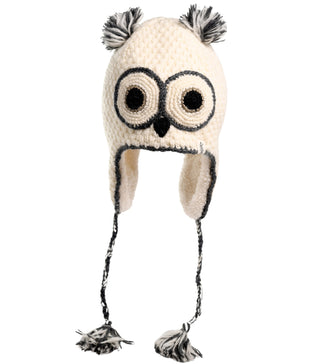 This Crochet Owl Hat is a hand-made, beige wool creation featuring large embroidered eyes, a small black beak, and gray pom-poms as ears. The hat has earflaps with braided strings that end in tassels.