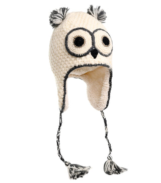 The Crochet Owl Hat is hand-made in white and showcases round eyes, a small beak, and ear tufts at the top. Crafted from wool, it includes ear flaps with braided tassels and black yarn outlining the owl's eyes and beak.