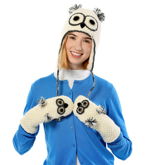 A person wearing the hand-made cream-colored Crochet Owl Hat, complete with pom-poms and matching mittens, poses against a white background. Dressed in a blue cardigan over a white top, they smile warmly at the camera.