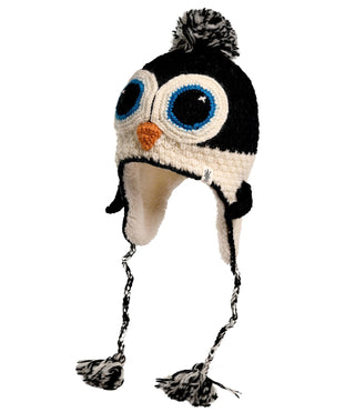 The Crochet Penguin Hat is hand-made in Nepal from high-quality wool. Designed with large blue eyes, an orange beak, and earflaps, it features braided tassels with black and white pom-poms at the ends. The hat is black and cream-colored with a black and white pom-pom on top.