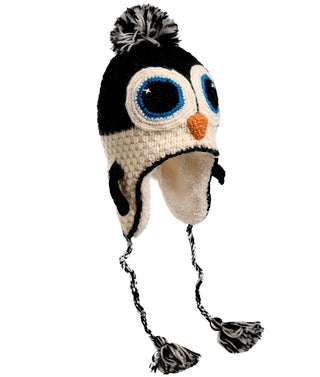 The Crochet Penguin Hat, hand-made in Nepal from high-quality wool, is a black and white hat designed to resemble a penguin face. It features large blue eyes, an orange beak, earflaps with braided tassels, and is topped with a charming black and white pom-pom.