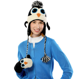 A woman wearing a blue cardigan and white shirt smiles while donning a hand-made Crochet Penguin Hat that resembles a cute penguin, complete with eyes, beak, and ear flaps. Made from high-quality wool in Nepal, the hat also features an adorable pom-pom on top.