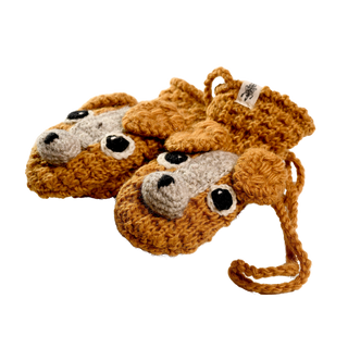 Introducing the Crochet Lucy Puppy Mittens, a charming pair of crocheted baby booties designed to resemble adorable dog faces. These handmade booties, crafted in Nepal, feature a primary orange-brown color with gray accents for the snouts and black buttons for the eyes. They are cleverly attached by a crocheted string, ensuring they stay together as a pair.