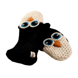 A pair of crochet penguin mittens featuring charming designs: one mitten is black with a white face, and the other is white with a black face. Both mittens have blue eyes and an orange beak. A small fabric tag is attached to one mitten, adding a touch of hand-made craftsmanship from Nepal.