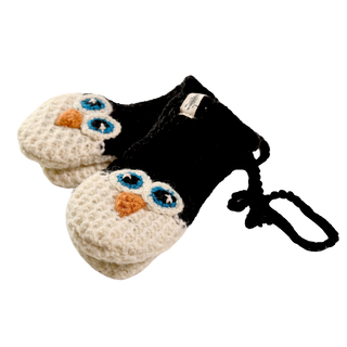 The Crochet Penguin Mittens are a pair of hand-made in Nepal knitted mittens designed to resemble penguin faces. The mittens are black with white sections for the penguins' faces, blue and black eyes, and orange beaks. They are connected by a black string and have a white tag attached to one mitten.