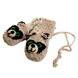 The Crochet Rockey Racoon Mittens feature two beige sherpa-lined knitted mittens connected by a string, adorned with raccoon faces that have green eyes and black and white detailing. Handmade in Nepal, they are displayed against a plain black background.