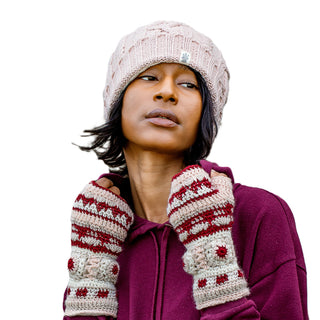 A person with short black hair, looking slightly to the side against a white background, is wearing a light pink knitted hat paired with Dreams Crochet Handwarmers. These fingerless gloves, handmade from 100% wool in Nepal, feature charming burgundy and white designs that complement their burgundy hoodie.