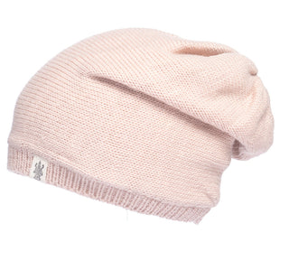 The Dekalb Slouch is a light pink, hand-knit beanie for cold weather with a small white tag featuring a gray logo on the folded edge. It’s displayed against a plain white background.