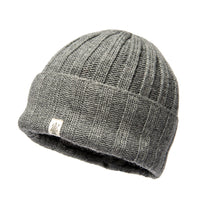 The Hand Loom Ribbed Beanie is a gray, ribbed knit hat crafted from merino wool, featuring a folded cuff and a small white logo tag, shown against a white background.