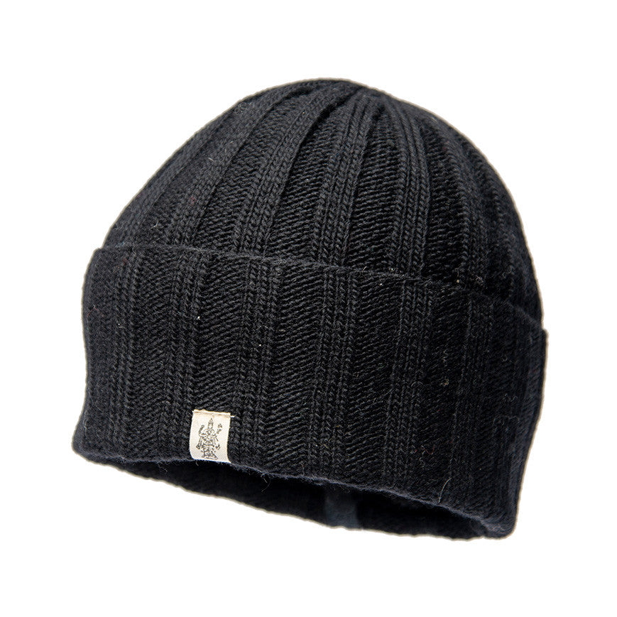 The Hand Loom Ribbed Beanie is a black merino wool hat featuring ribbed knit construction, a folded brim, and a small rectangular fabric tag with a graphic logo on the front. The background is plain white.