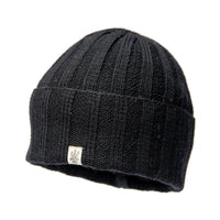 The Hand Loom Ribbed Beanie is a black merino wool hat featuring ribbed knit construction, a folded brim, and a small rectangular fabric tag with a graphic logo on the front. The background is plain white.
