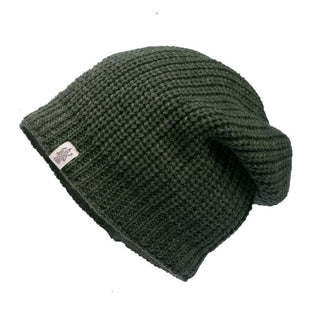 The Cardigan Knit Slouch, a dark green knit beanie with a ribbed texture and a slight slouch, showcases its handmade in Nepal craftsmanship against a white background. The beanie features a small white tag with the brand's logo sewn into the lower edge.