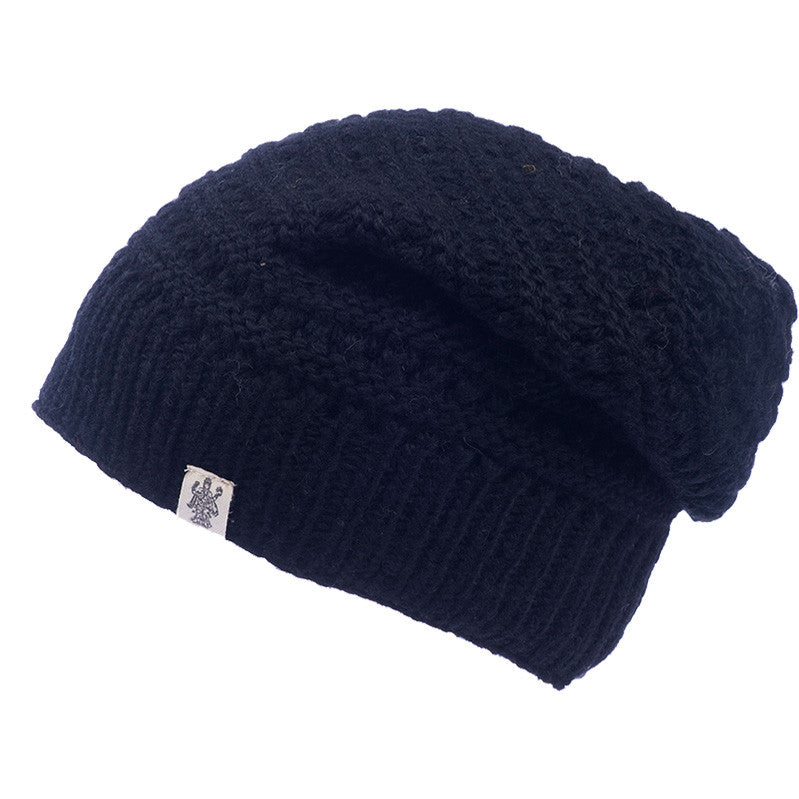 The Hand Knit Boho Slouch is a black, handmade wool beanie hat featuring a ribbed brim and a small white rectangular tag with a logo on the side. Displayed against a plain white background, this slouch-style hat combines artisanal craftsmanship with modern design.