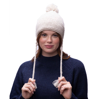 Sentence with product name: A person wearing a cream-colored, wool knitted beanie with a pom-pom and Verona earflap tassels, paired with a navy blue sweater, gently holds the Verona earflap tassels.