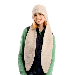 A person with shoulder-length blonde hair is wearing an oversized ear beanie in beige, featuring long scarf-like extensions. They are smiling and dressed in a green sweater with a black puffer vest against a solid black background.