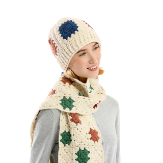 A person wearing the Crochet Patch Hat and a matching scarf adorned with a floral pattern. Both the hat and scarf feature a cream color with blue, green, and red flower motifs. The person is looking slightly to the side and dressed in a light gray top, set against a plain white background.