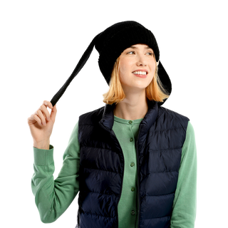 A person with shoulder-length blond hair is wearing a black Rib EarPup Hat, a green long-sleeve shirt, and a dark blue puffer vest. They are holding the end of their beanie to the side and smiling, with their eyes looking slightly off-camera.