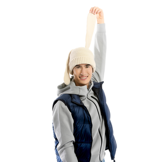 A person wearing a Rib EarPup Hat in white, along with a gray hoodie and navy blue puffer vest, smiles while holding up the end of their hat with one hand. The background is transparent.