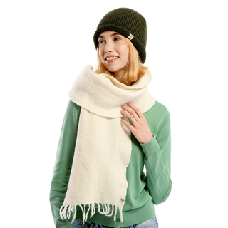 A person wearing a Baxter Hat winter beanie in green, a light green sweater, and a large white Sherpa fleece scarf. The person has short, light-colored hair and is smiling with a relaxed expression, with their right hand adjusting the scarf. The background is plain white.