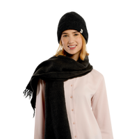 A woman wearing the Hand Knit Bond Hat in black along with a large black scarf and a pale pink button-up shirt stands against a plain white background, smiling gently at the camera.