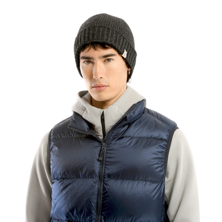 A person is wearing the black Cabrini Hat with a cozy fleece lining, a light gray hoodie, and a dark blue puffer vest. They are looking directly at the camera against a solid black background.