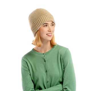 A person with shoulder-length blonde hair is wearing the Cabrini Hat, a beige knit beanie with a subtle knit texture, paired with a light green button-up cardigan. They are looking slightly to the left with a relaxed smile against a plain black background.