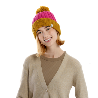 A person wearing a Cherry Pom Hat, a beige cardigan, and a brown ribbed shirt smiles at the camera against a plain black background.