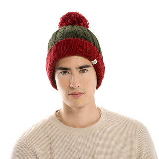 A person is wearing a Cherry Pom Hat, featuring red and dark green knit design with a red pom-pom on top. They are also dressed in a beige waffle-knit long-sleeve shirt and are looking straight ahead with a neutral expression. The background is plain black.