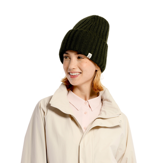 A person with a smiling expression is wearing a dark green Cooper Hat, a winter essential, and a light beige coat over a light pink collared shirt. The background is black.