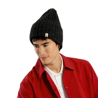 A person wearing a stylish outdoor headwear—a black Cooper Hat made of premium merino wool—pairs it with a white polo shirt and a red jacket, looking downward to the left. The background is plain black.